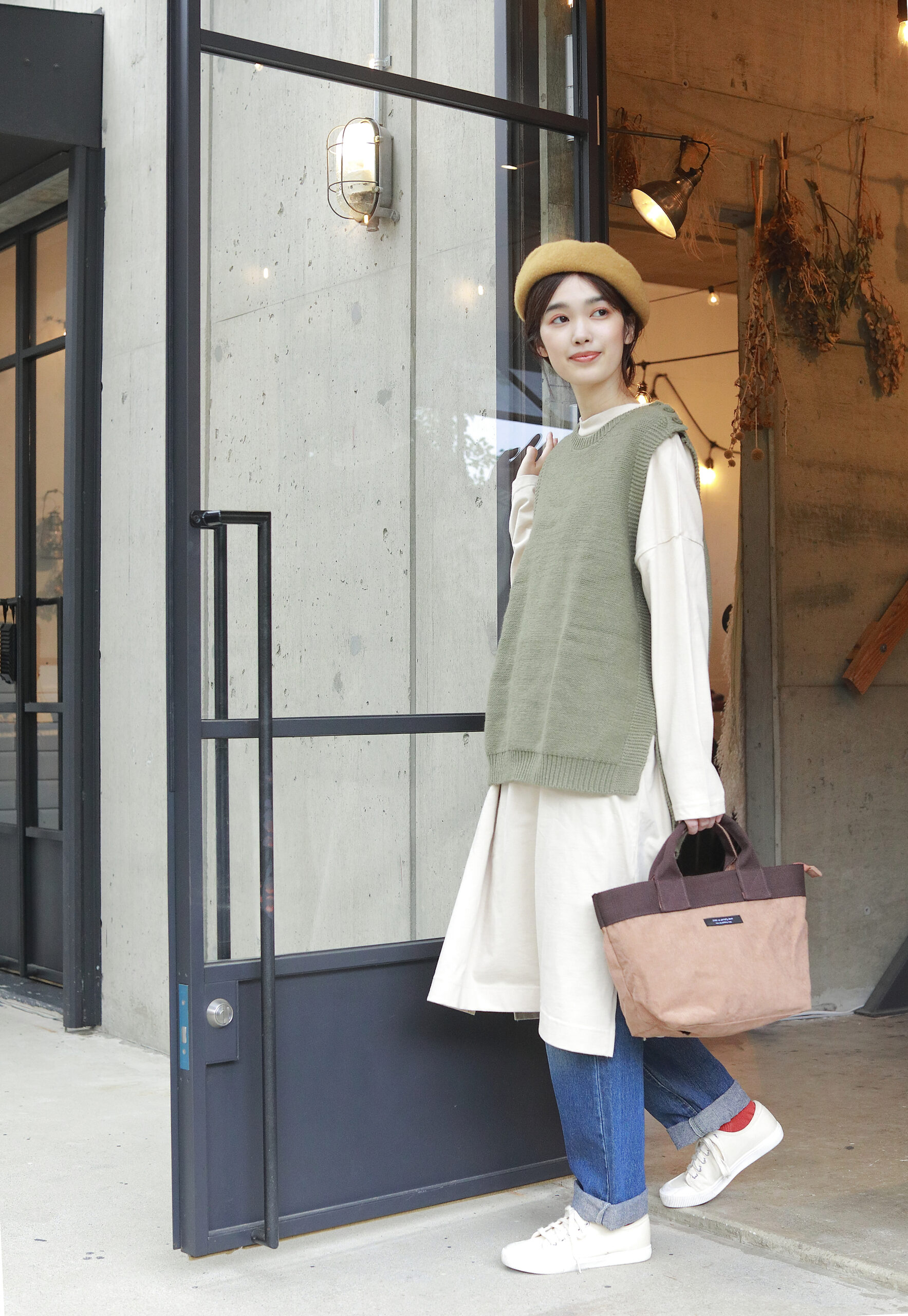 黄色いベレー帽をかぶってカーキのベストをロングのTシャツと合わせた、女性モデルがお店から出てくる写真