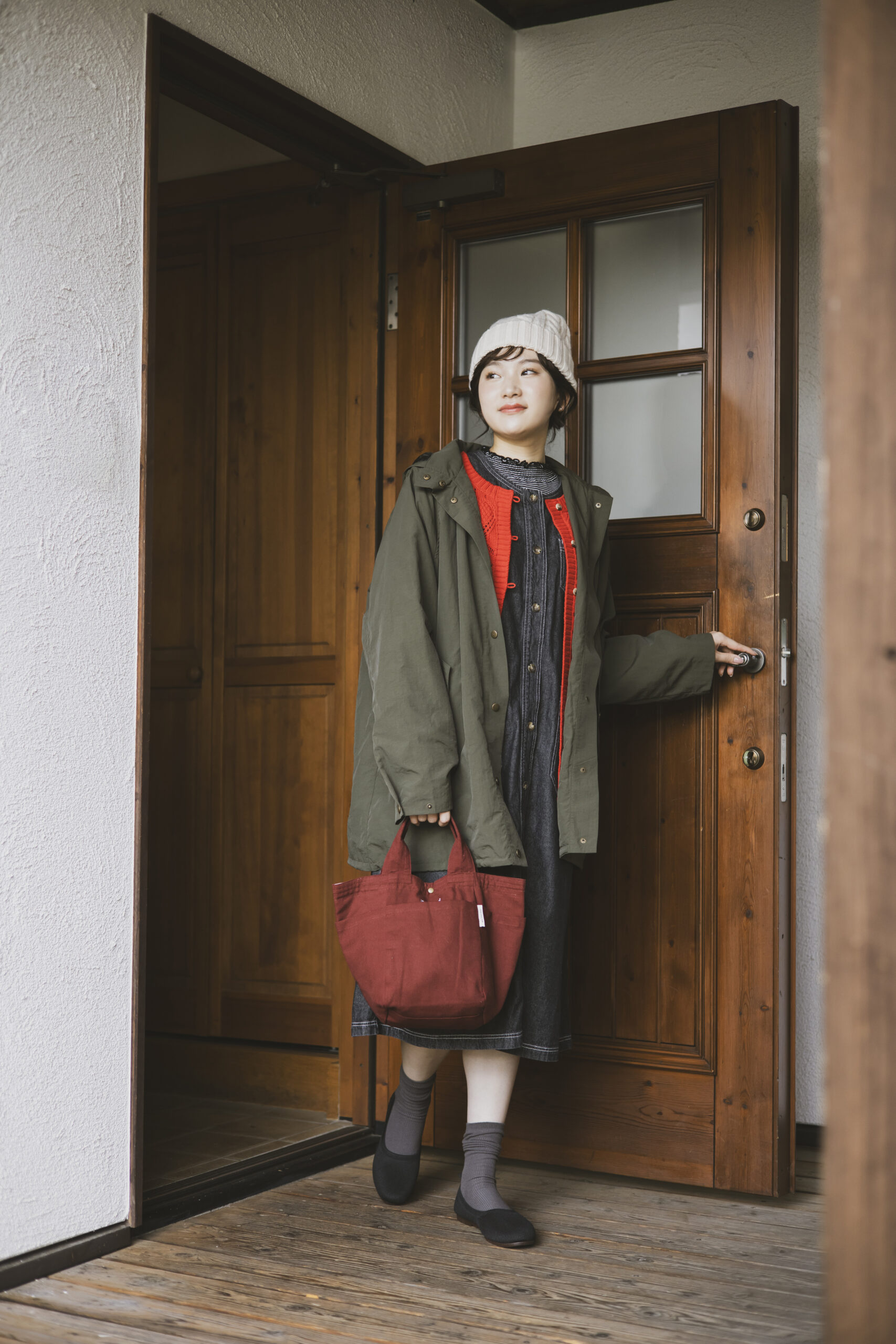 カーキのコートの中に赤いカーディガンとデニムのワンピースを纏い、アイボリーのニット帽を被って手にバーガンディ色のバッグを持っている女性