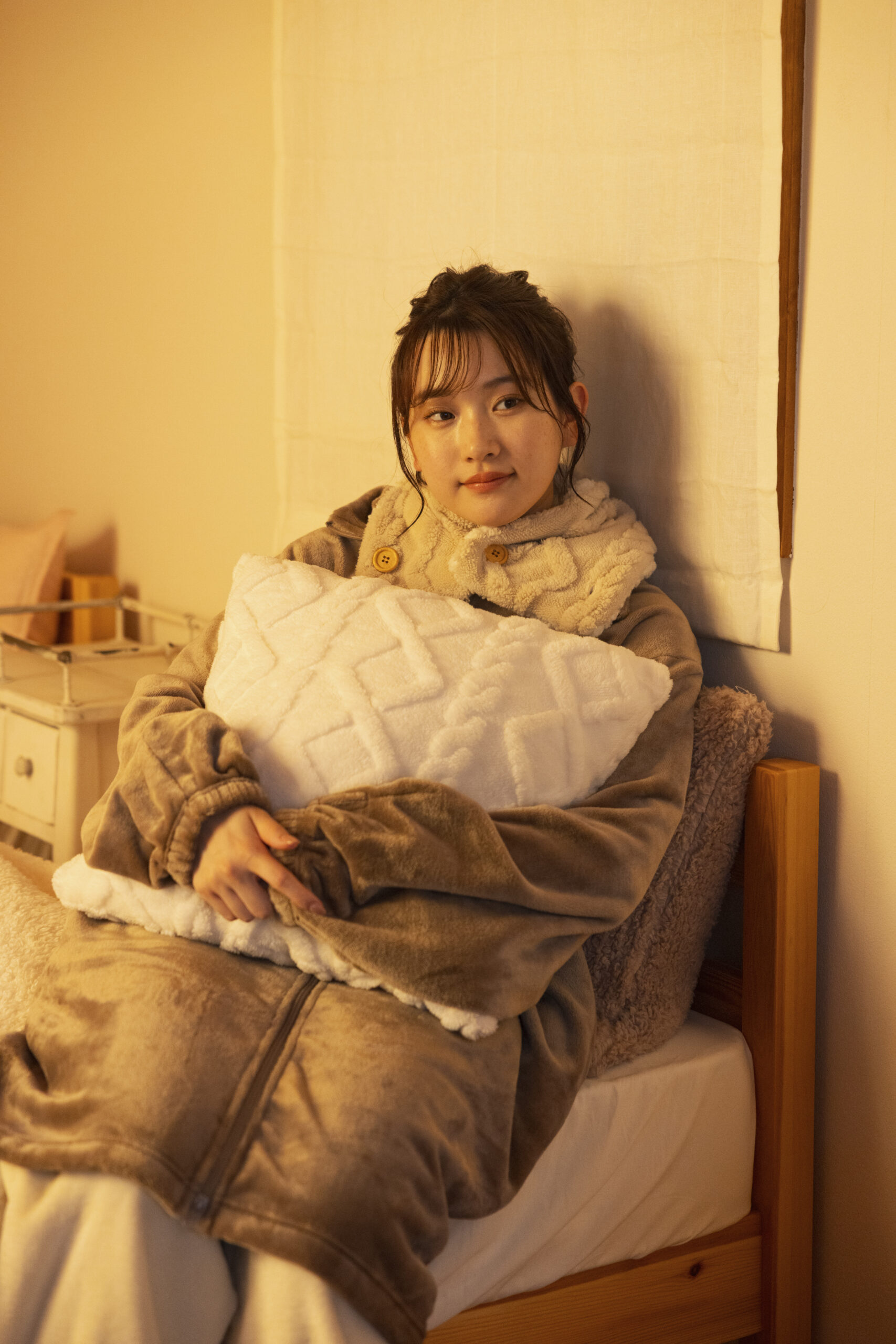 クッションをかけてベッドに腰掛ける、首にボアティペットを巻きブラウンの暖かそうなリラックスガウンジャケットを纏った女性