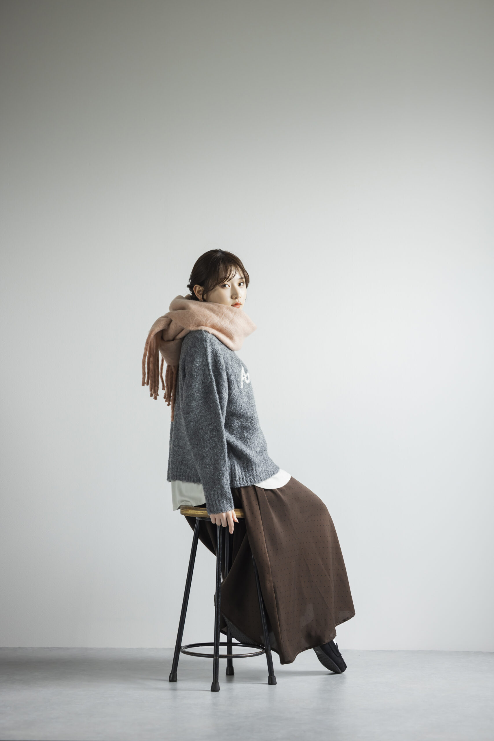 ピンクのマフラーを巻き、グレーのニットにブラウンのスカートをはいて椅子に座っている女性