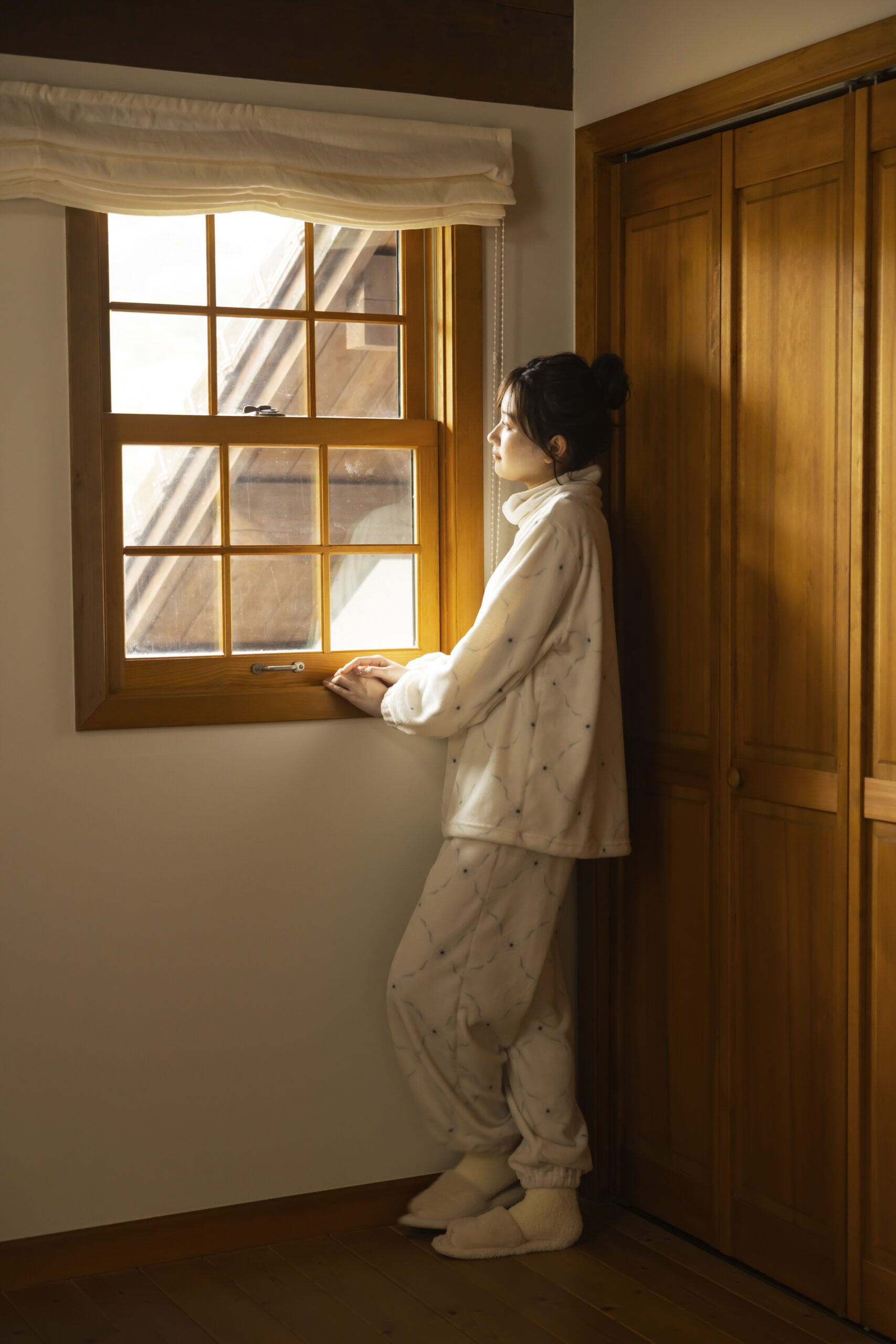 白ベースの優しい生地感のルームウェアを身にまとい、窓の外を眺めている女性