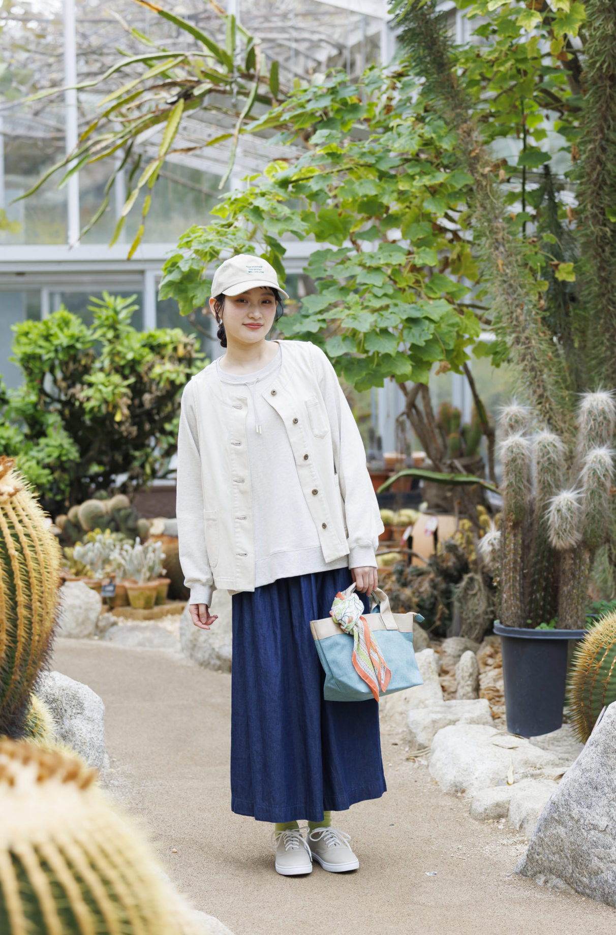 アイボリーのキャップをかぶり、アイボリーのトップスにアイボリーのデニムベストとデニムロングスカートを履いて水色とベージュの切り替えのあるバッグにスカーフを付けたバッグを手に持った女性の画像。