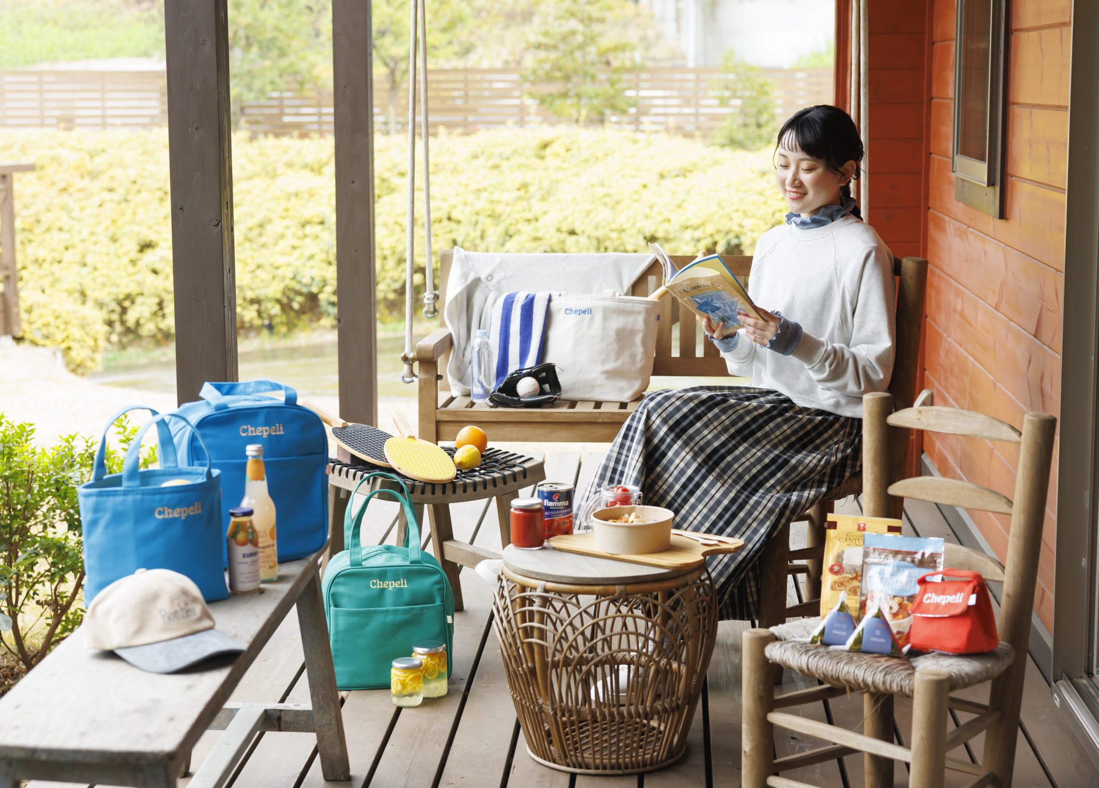 ラケットやボール、おにぎりや弁当やジュースや瓶と帽子や色々なカラーや形のランチボックスに囲まれてグレーのトップスとチェックのロングスカートを履いて本を読んでいる女性の画像