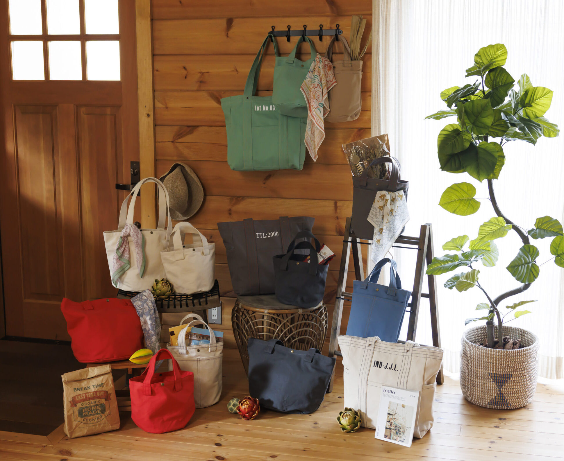 植物や本やスカーフや紙袋と赤、青、紺、白、緑、ブラウンの色の大きさがバラバラのバッグが置いている画像