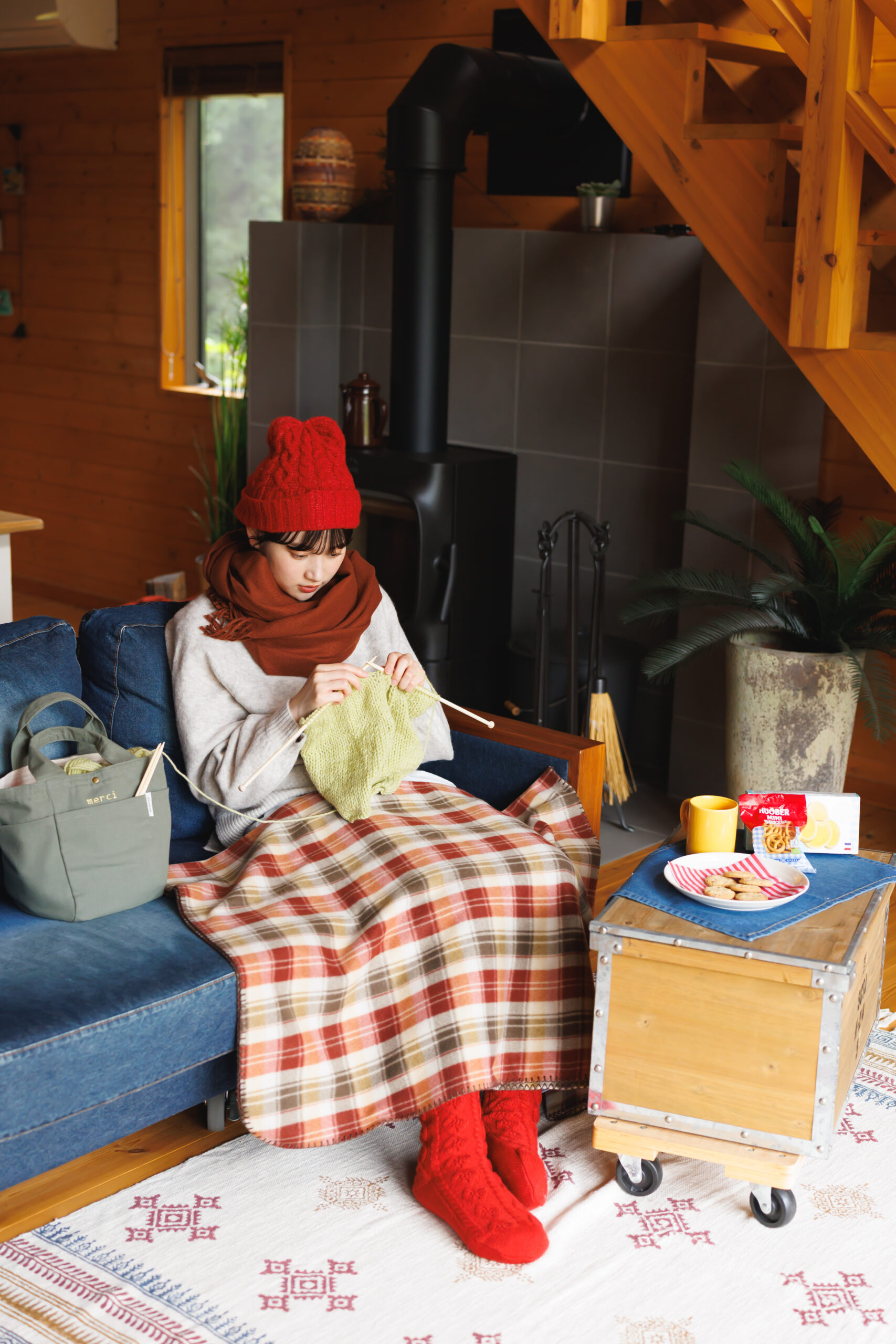 赤の帽子に茶色いマフラー、アイボリーのニットにチェックのブランケットを足にかけ赤い編み柄ソックスを履いて編み物をしてくつろいでいる女性の画像。