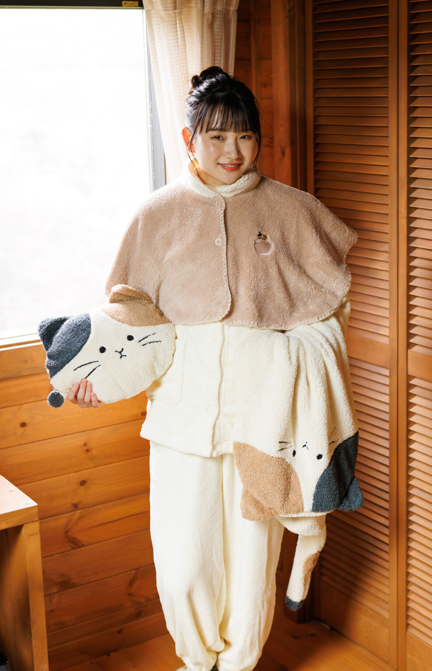 白いパジャマの上にベージュのポンチョを羽織ってネコモチーフのクッションとブランケットを持っている女性