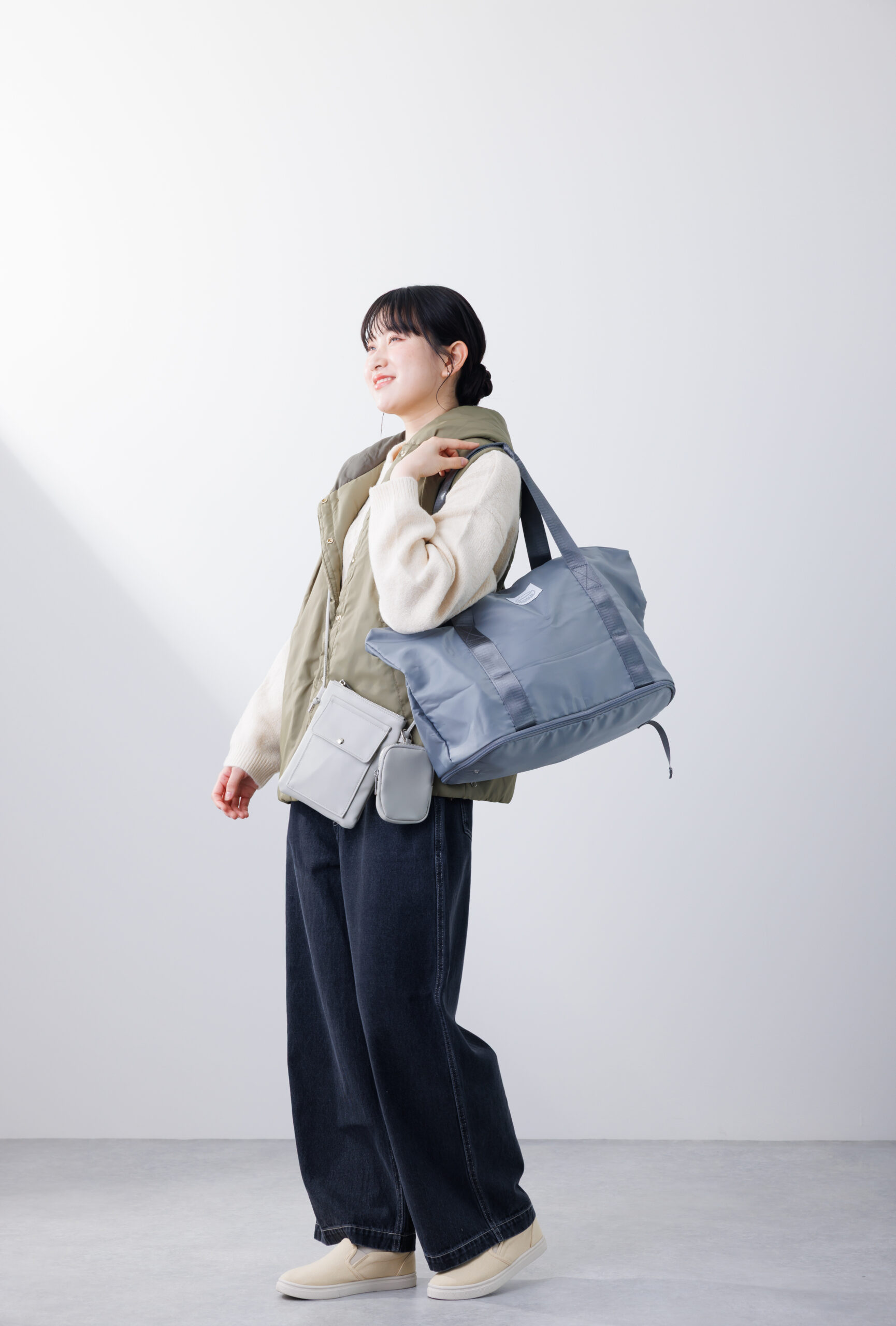 カーキのベストを羽織った女性がブルーグレーのボストンバッグとグレーのショルダーバッグを持って出かけている画像