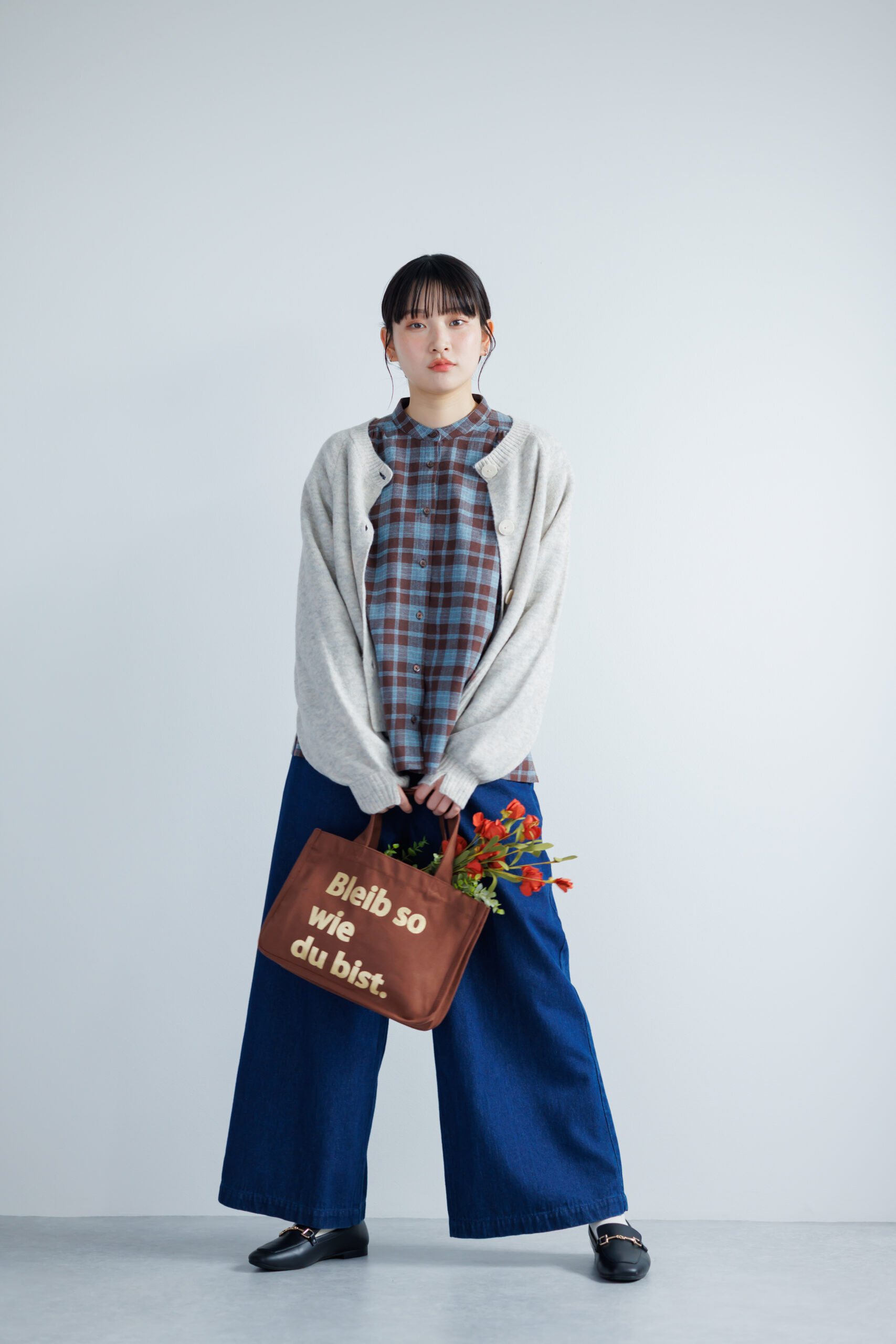 ブルーとブラウンのチェック柄カットソーにグレーのカーディガンを羽織ってブルーのワイドパンツを履いて、ブラウンのロゴバッグを持っている女性